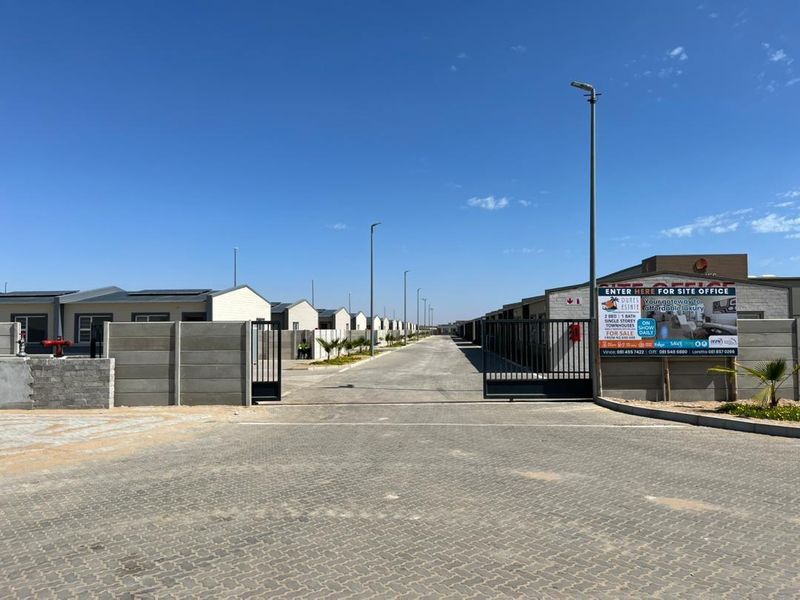 View of the entrance and paved access road within a modern townhouse estate, featuring security gates and perimeter walling.