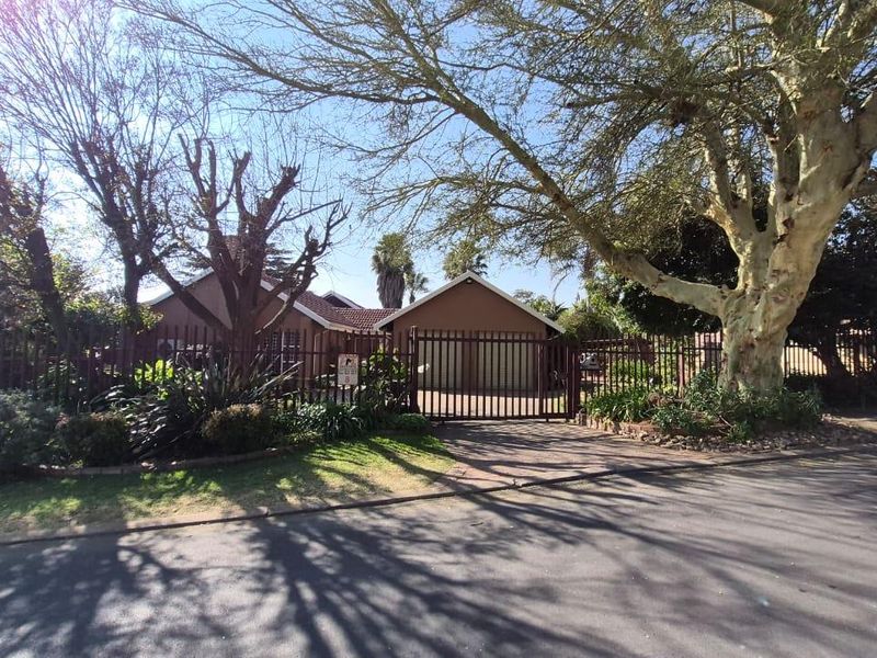 Front view of the property featuring a paved driveway, secure metal gate, and established trees providing shade to the building facade.