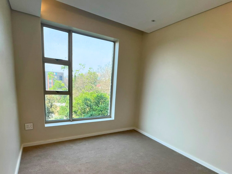 A bright, empty bedroom featuring neutral walls, carpet flooring, and a large window providing natural light and an exterior view.