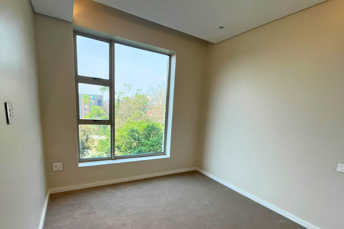 A bright, empty bedroom featuring neutral walls, carpet flooring, and a large window providing natural light and an exterior view.