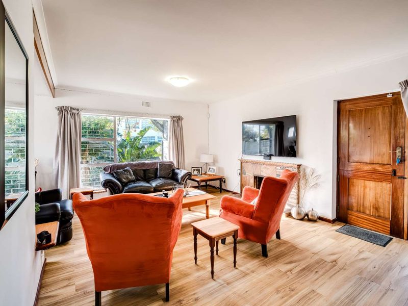 A bright living room featuring timber-look flooring, a fireplace, and a solid timber entrance door with natural light from a large window.