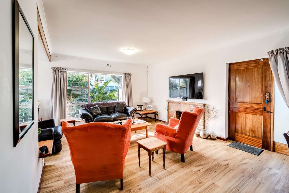 A bright living room featuring timber-look flooring, a fireplace, and a solid timber entrance door with natural light from a large window.
