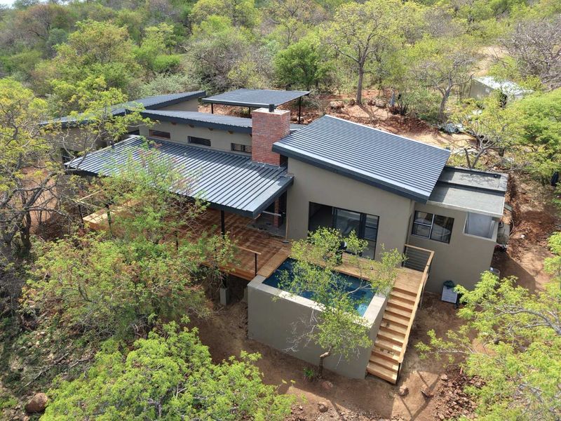 Aerial view of a modern house featuring a metal roof, brick chimney, wooden deck, and an integrated swimming pool set in a natural landscape.