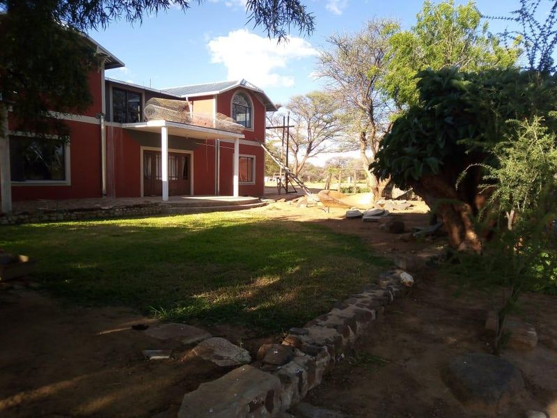 Rear view of a two-story farmhouse featuring a covered patio, balcony, and stone retaining wall, set within a large, partially landscaped garden area.
