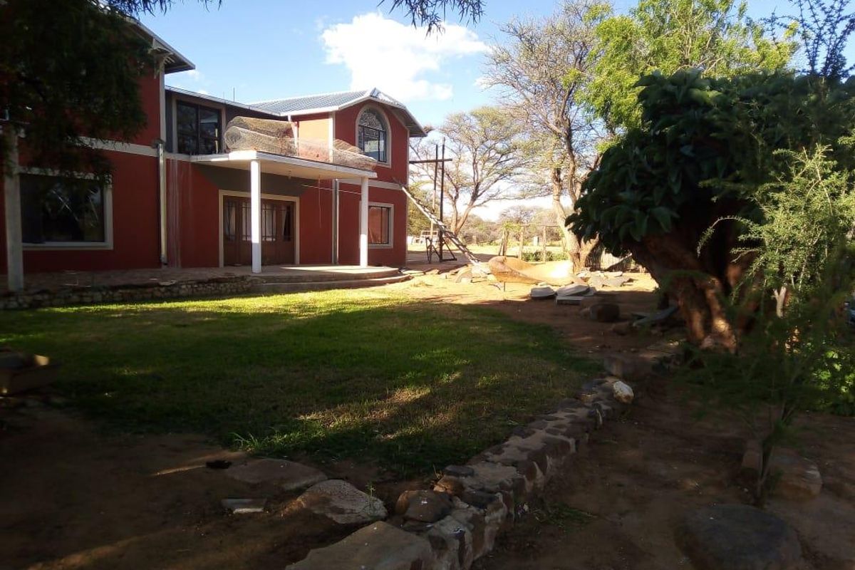 Rear view of a two-story farmhouse featuring a covered patio, balcony, and stone retaining wall, set within a large, partially landscaped garden area.