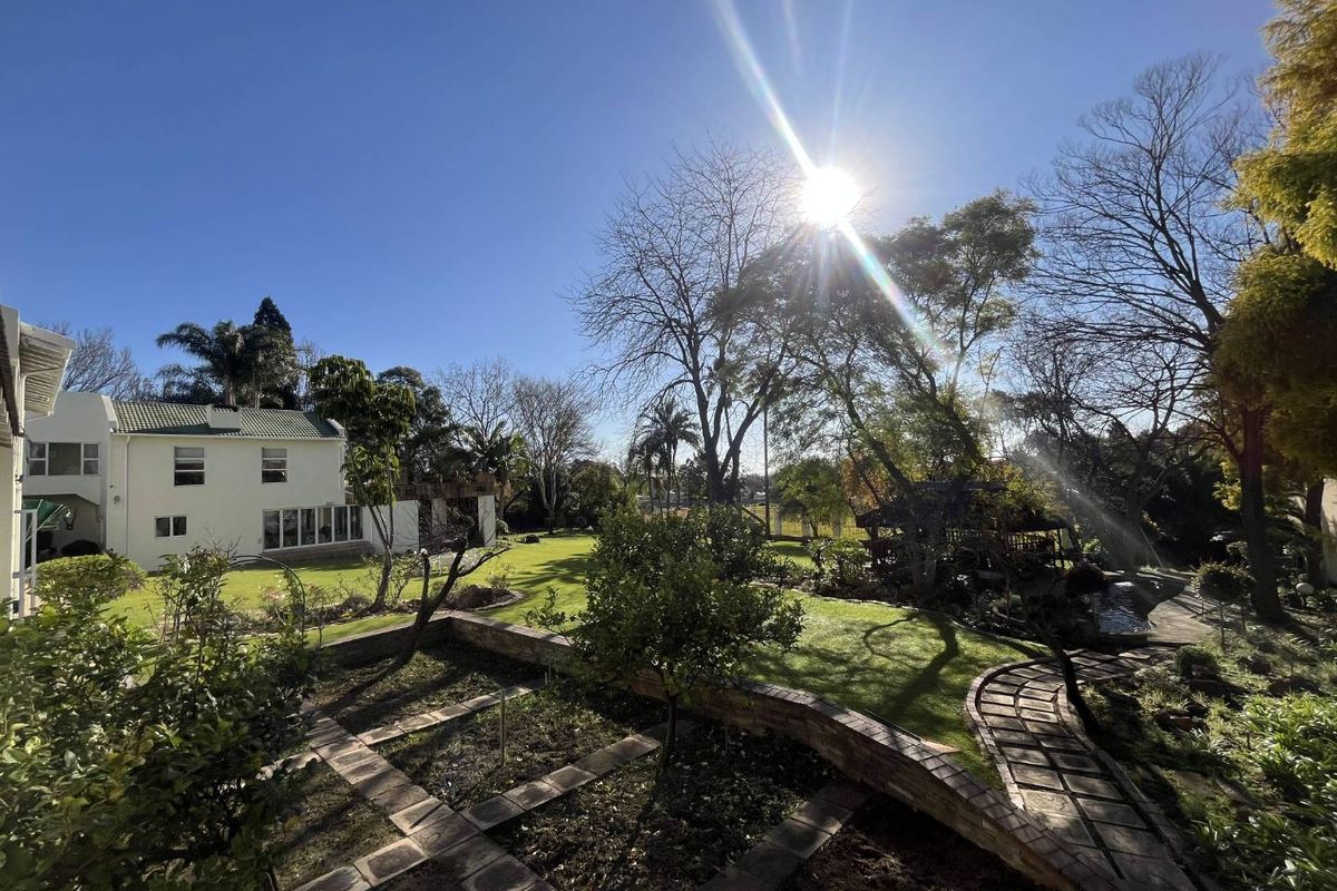 A spacious rear garden featuring manicured lawns, established trees, brick-edged garden beds, and a paved pathway leading through the landscaped grounds.