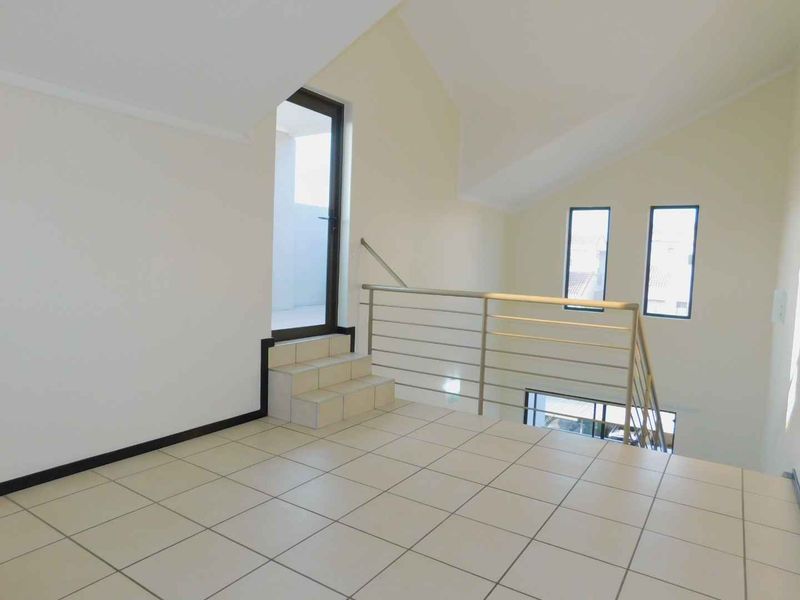 An empty, light-filled living area featuring tiled floors, a vaulted ceiling, and a metal balustrade overlooking the lower level.