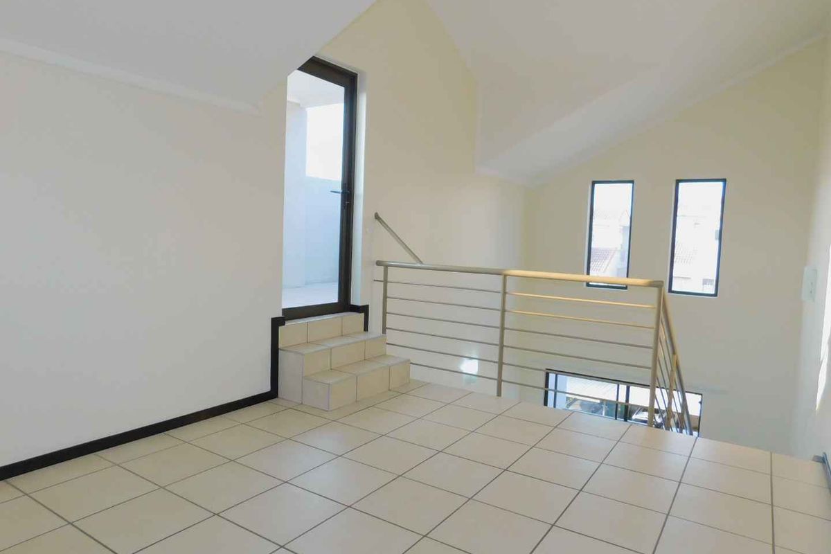An empty, light-filled living area featuring tiled floors, a vaulted ceiling, and a metal balustrade overlooking the lower level.
