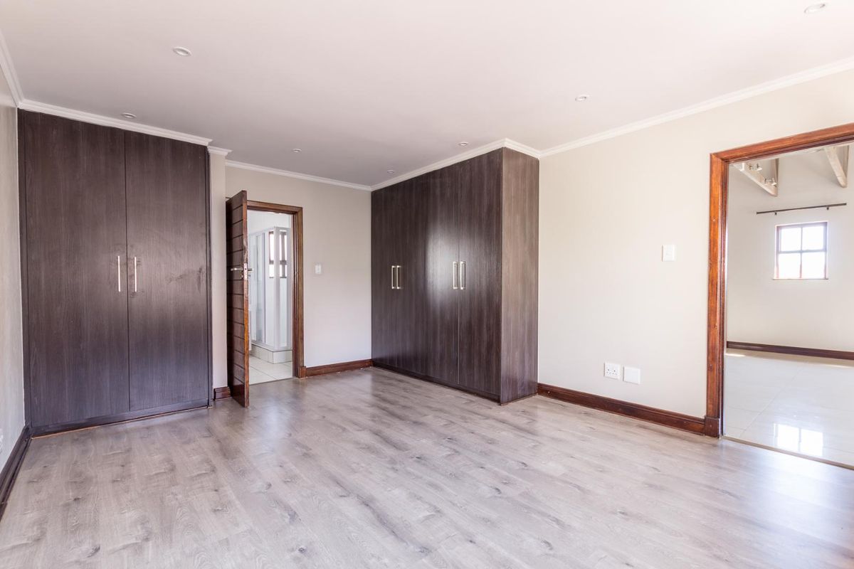 A spacious bedroom featuring built-in dark wood cabinetry, light wood-look laminate flooring, and recessed ceiling lighting, with an adjoining en-suite bathroom.