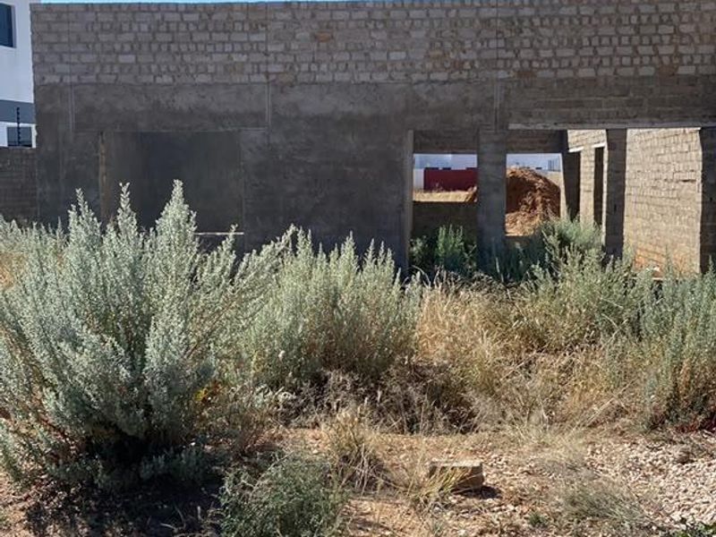 An unfinished masonry structure showing the exterior shell of a property under construction, set on a plot with overgrown vegetation.