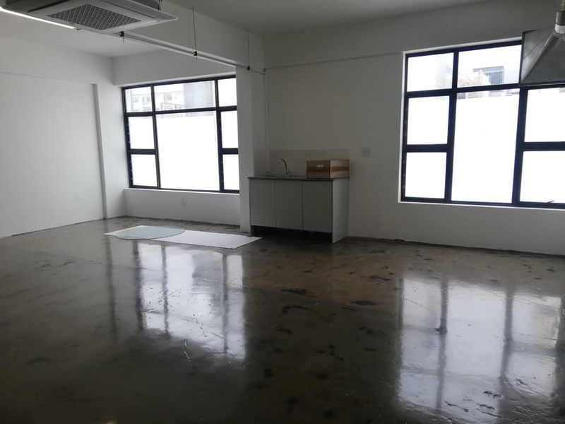An open-plan office space featuring polished concrete floors, large black-framed windows, and a compact kitchenette area with a sink.