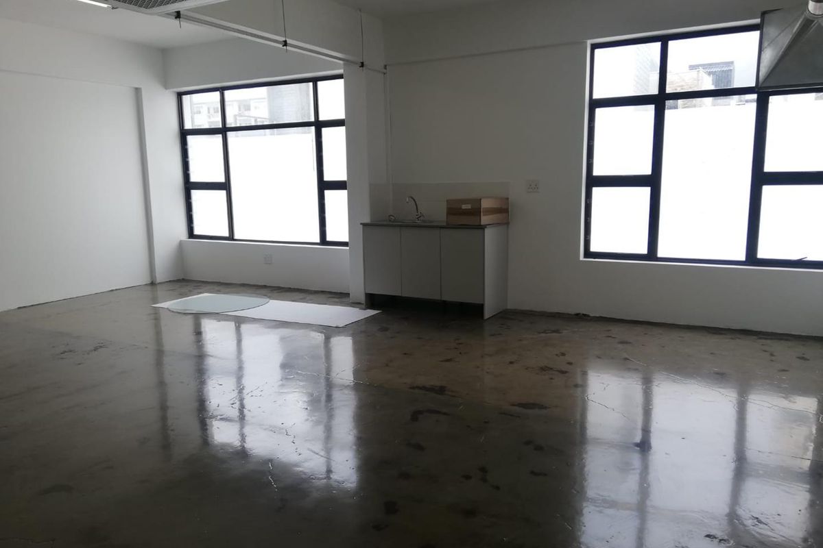 An open-plan office space featuring polished concrete floors, large black-framed windows, and a compact kitchenette area with a sink.