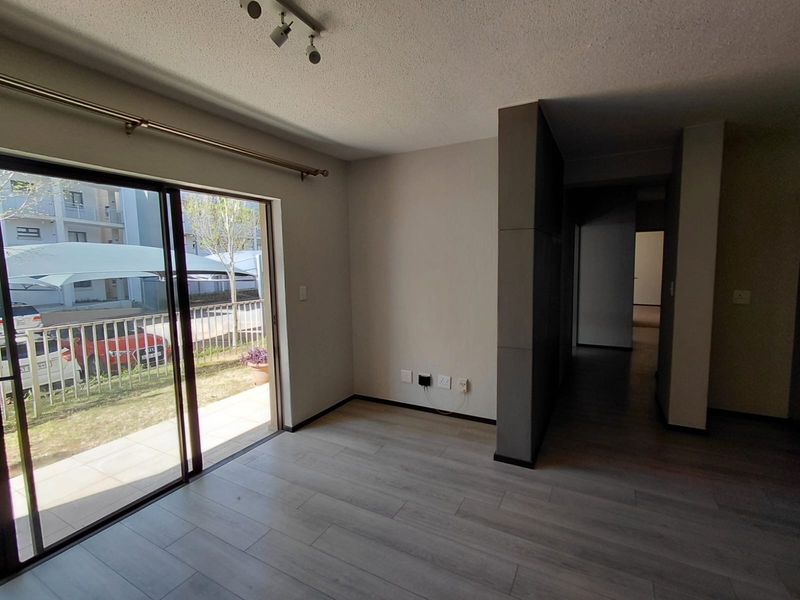 An empty, well-lit living room featuring modern laminate flooring, a large sliding glass door leading to a patio, and neutral wall finishes.