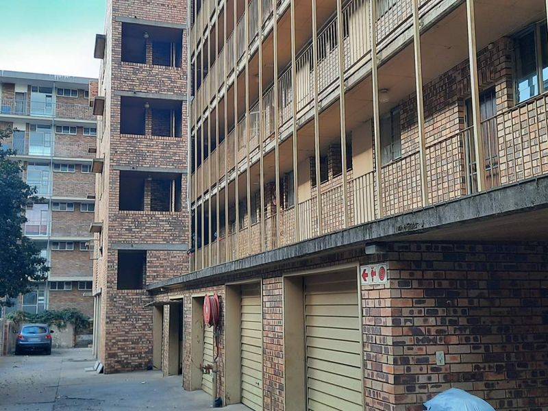 Exterior view of a multi-story brick apartment building featuring ground-level garages, open balconies with metal railings, and a paved driveway area.