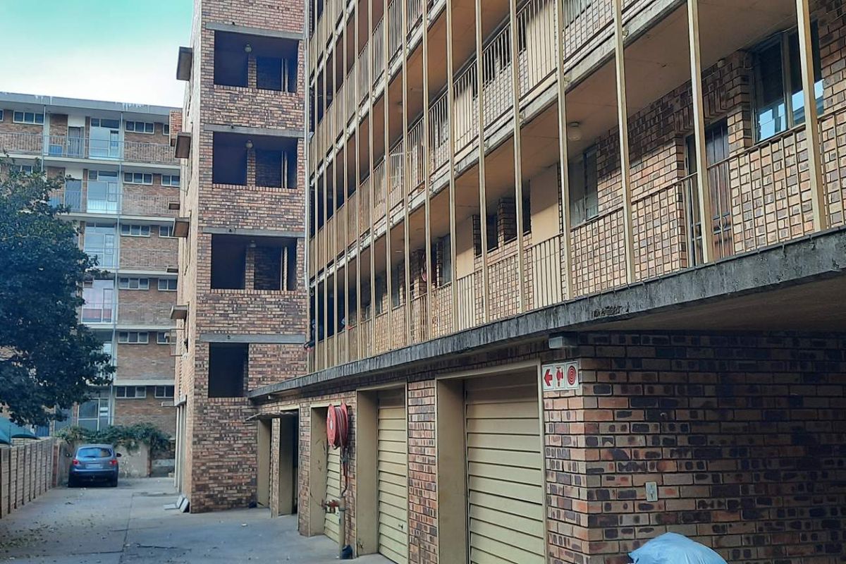 Exterior view of a multi-story brick apartment building featuring ground-level garages, open balconies with metal railings, and a paved driveway area.