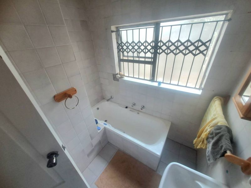 A functional bathroom featuring a built-in bathtub, tiled walls and flooring, and a window fitted with a security gate.