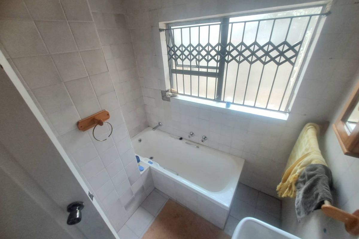 A functional bathroom featuring a built-in bathtub, tiled walls and flooring, and a window fitted with a security gate.