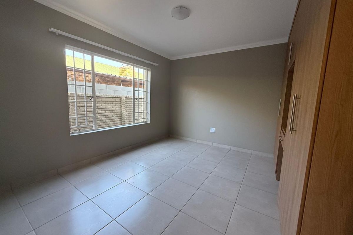 A bright, empty bedroom featuring neutral tiled flooring, built-in wooden cabinetry, and a large window with security bars.