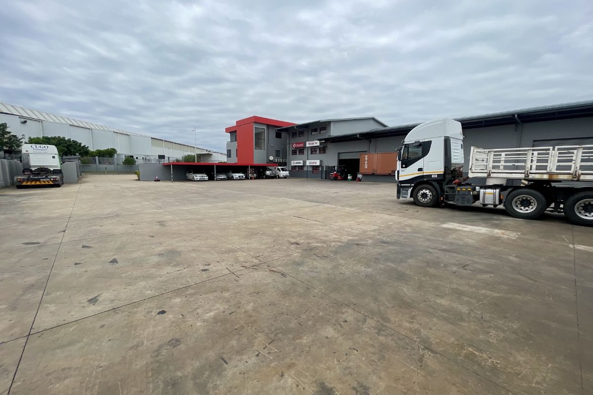 Spacious industrial yard featuring a large concrete surface, warehouse facility, and covered parking area suitable for heavy vehicle access and logistics operations.