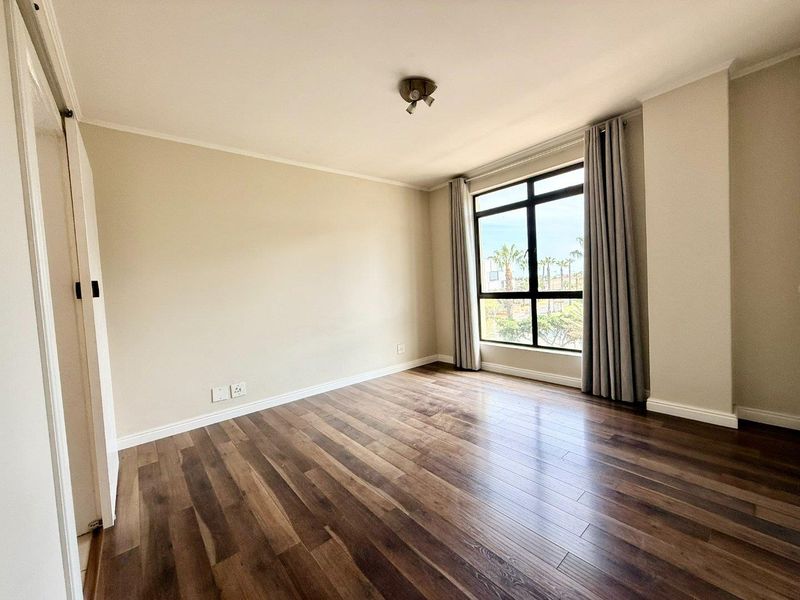 A bright, empty bedroom featuring modern laminate flooring, a built-in cupboard, and a large window providing natural light.