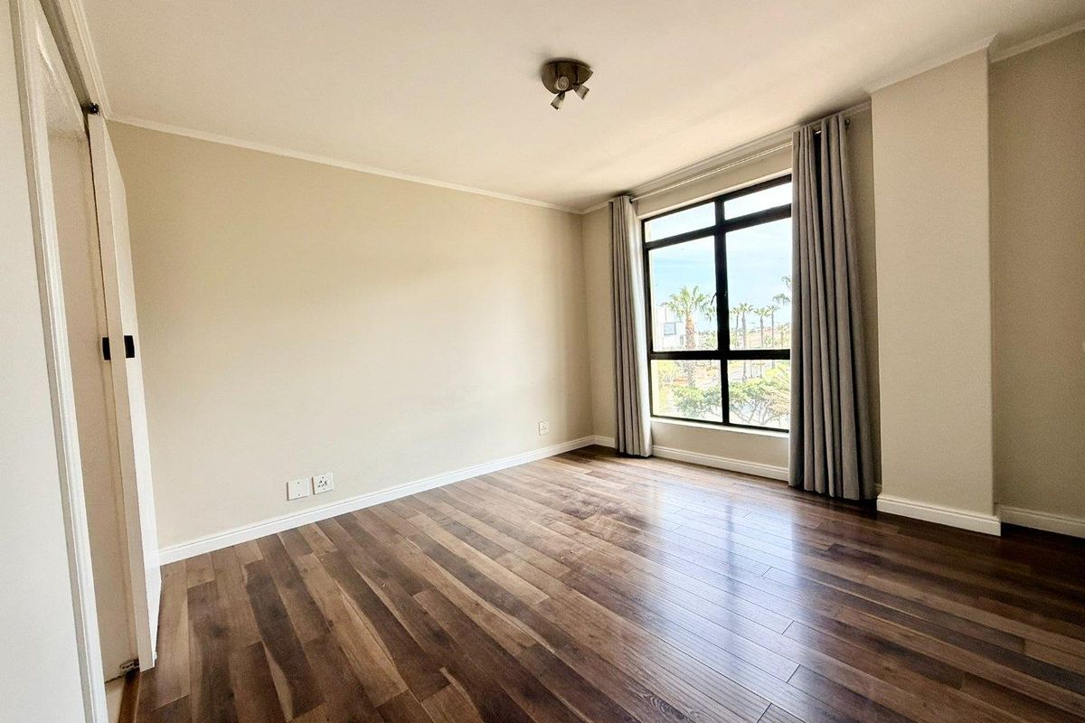 A bright, empty bedroom featuring modern laminate flooring, a built-in cupboard, and a large window providing natural light.