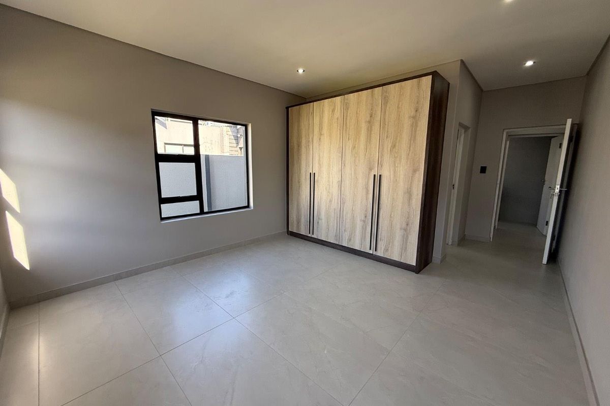 A bright, modern bedroom featuring large format floor tiles, recessed downlighting, and a built-in wardrobe with a wood-grain finish.