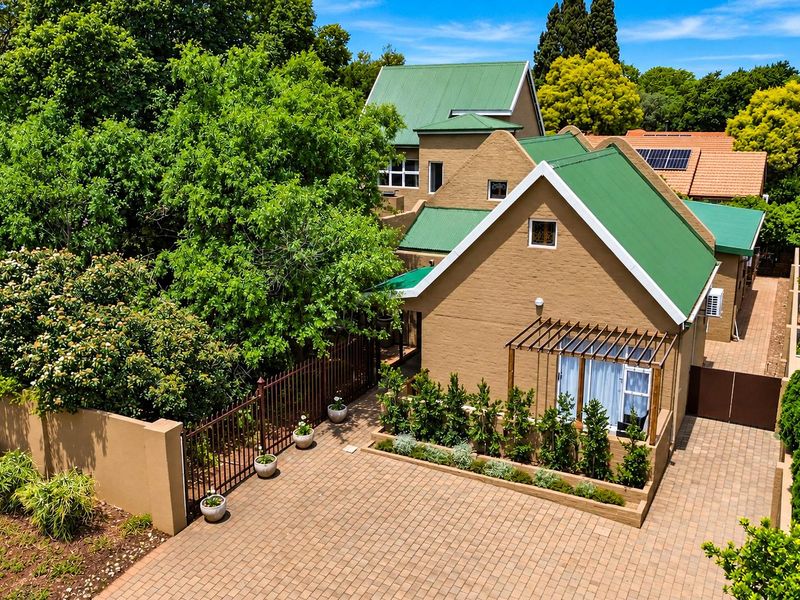 Aerial view of a residential property featuring a green metal roof, brick exterior, paved driveway, and established garden landscaping.