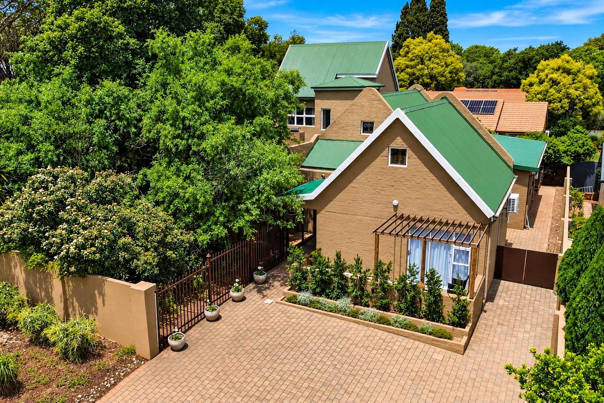 Aerial view of a residential property featuring a green metal roof, brick exterior, paved driveway, and established garden landscaping.
