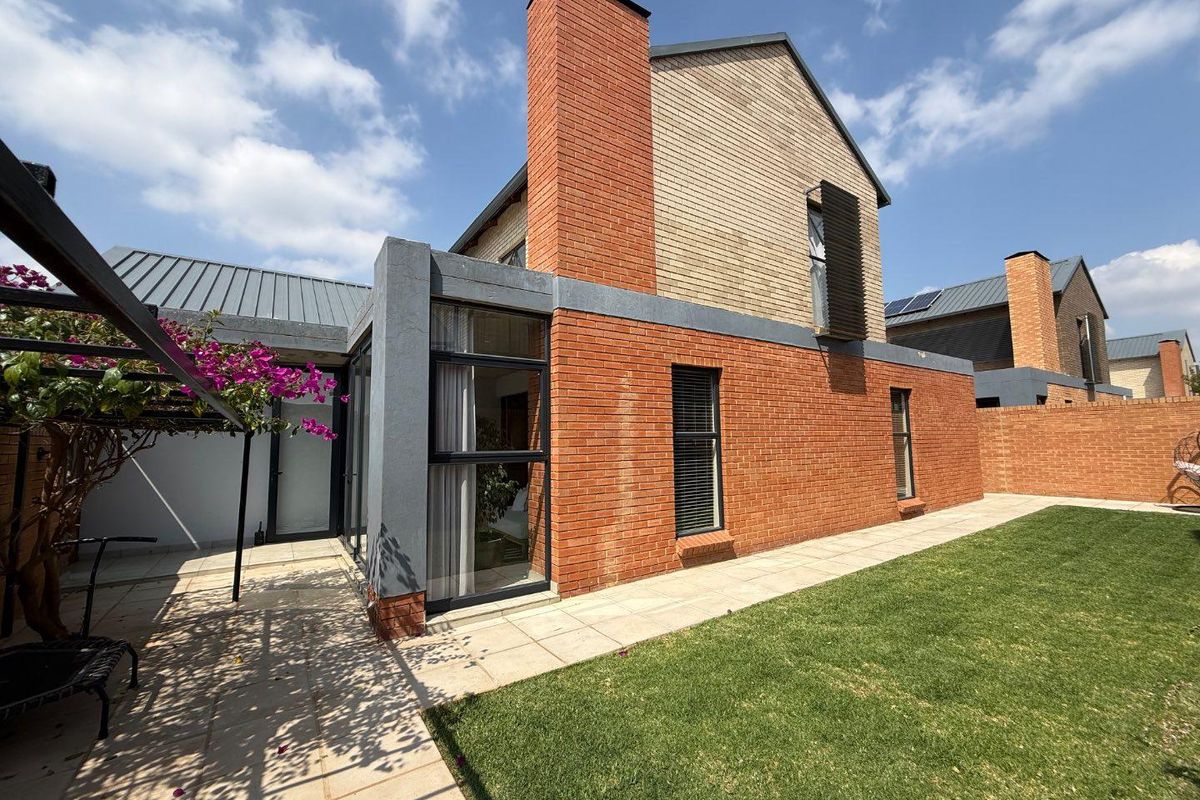 Rear view of the townhouse featuring a brick facade, prominent chimney, tiled patio area, and a well-maintained lawn.