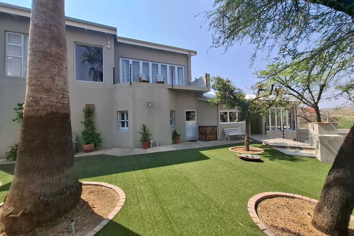 Rear view of the property featuring a landscaped garden with artificial turf, mature trees, and a raised balcony area accessible from the upper level.
