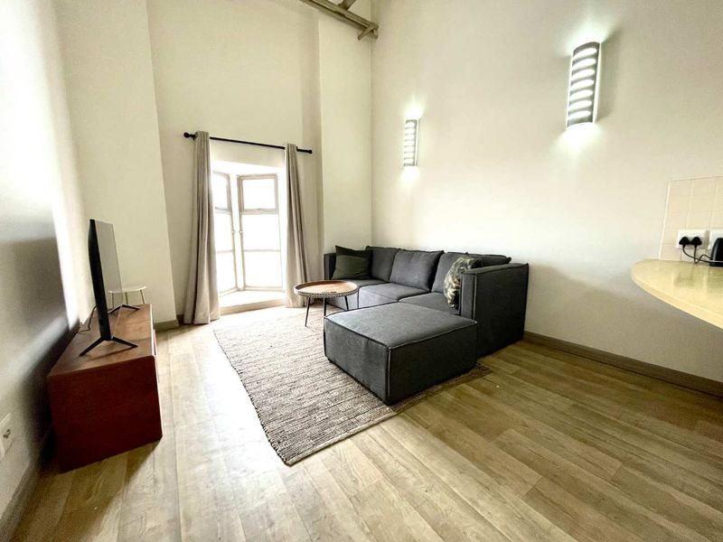 A bright living room featuring high ceilings with an exposed beam, laminate flooring, wall-mounted lighting, and a bay window.