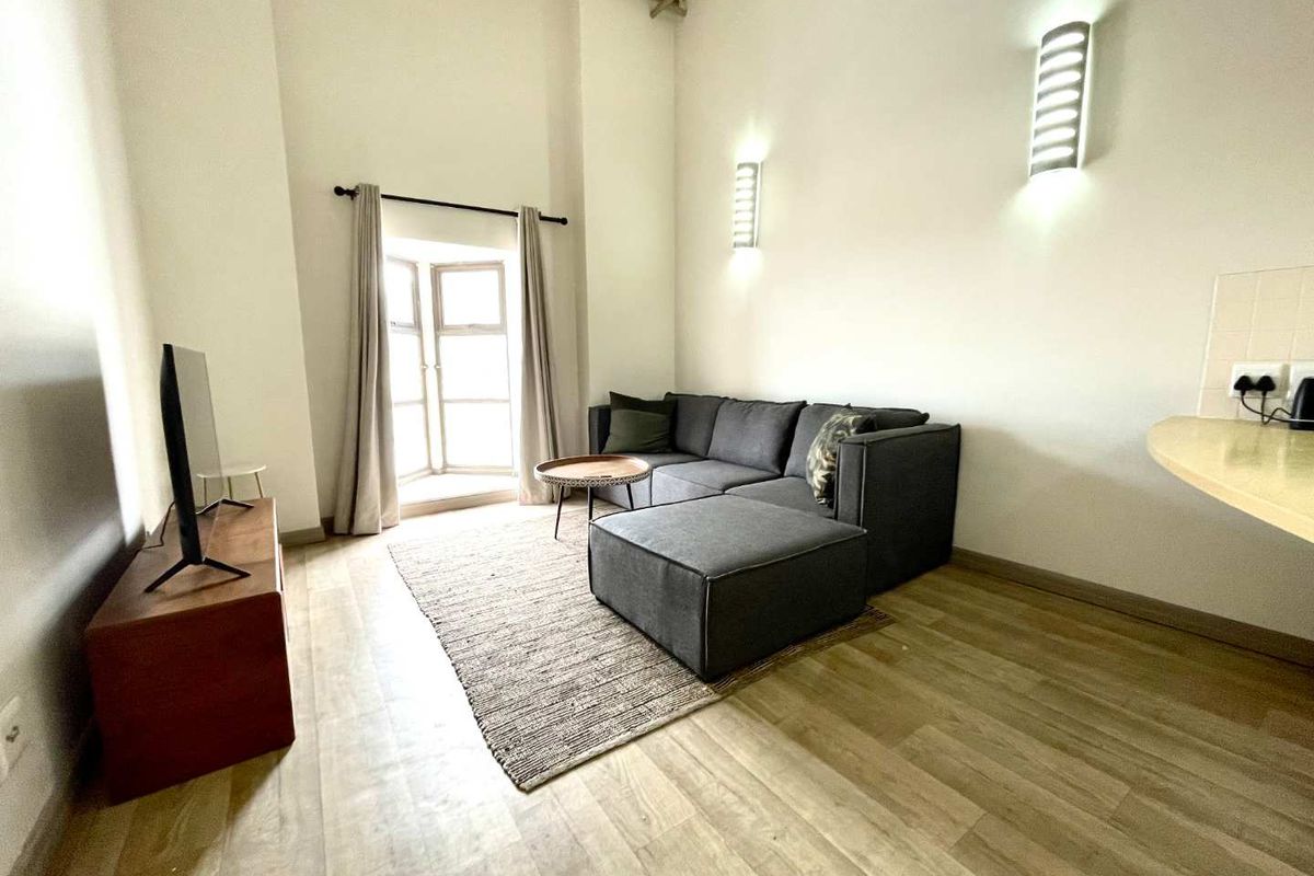 A bright living room featuring high ceilings with an exposed beam, laminate flooring, wall-mounted lighting, and a bay window.