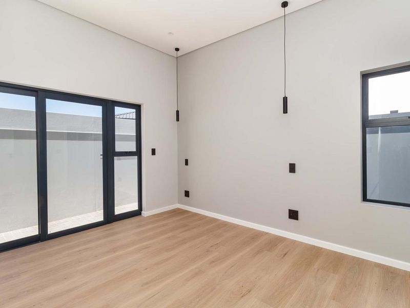 A bright, modern, and empty bedroom featuring light wood-look laminate flooring, sleek black aluminium window and door frames, and contemporary pendant lighting.