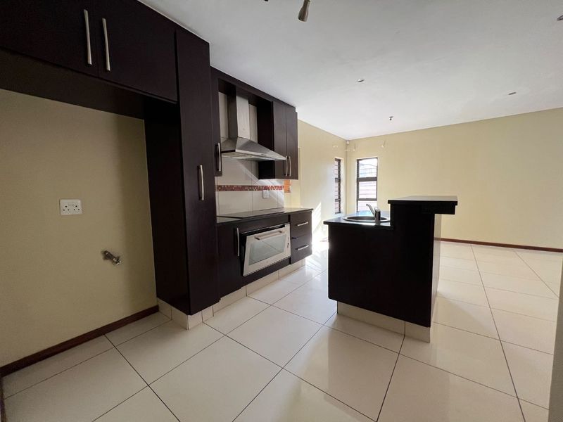 A modern, empty kitchen featuring dark wood cabinetry, a built-in oven and hob, a central island with a sink, and light-colored floor tiles.
