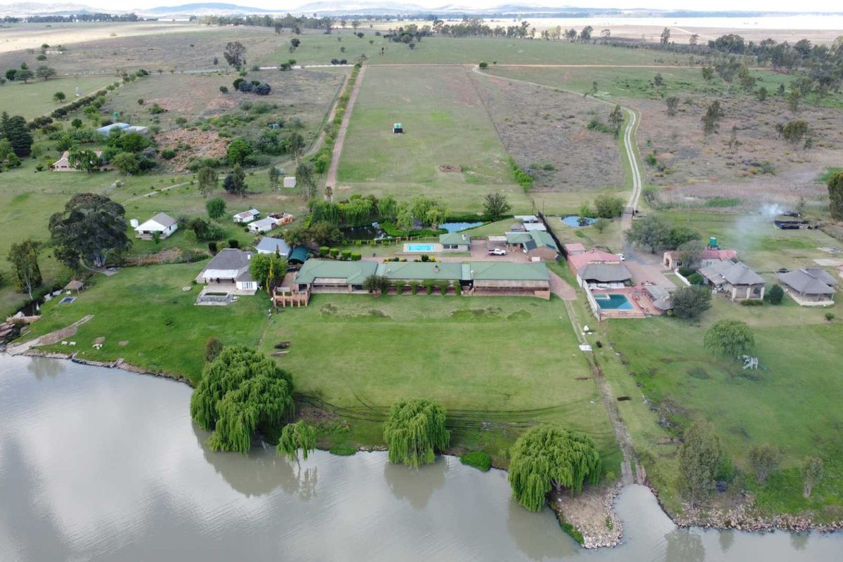 Aerial view of a large rural property featuring multiple buildings, swimming pools, expansive lawns, and direct access to a dam.