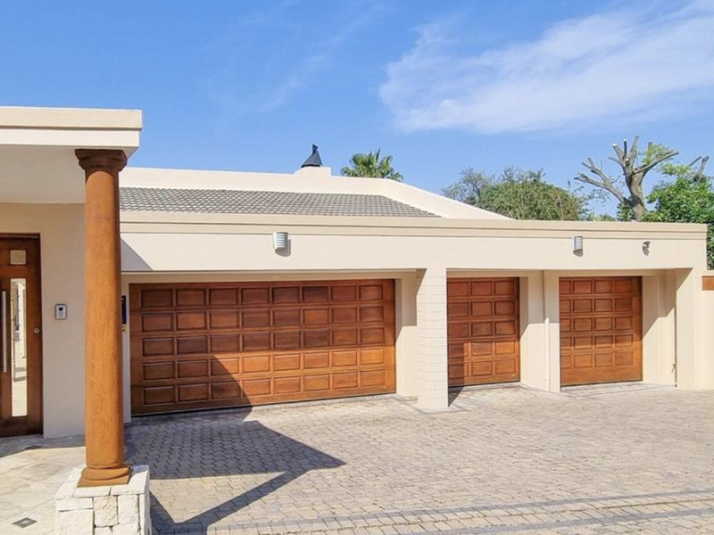 Front elevation featuring a triple garage with wooden doors, a paved driveway, and a covered entrance with a decorative pillar.