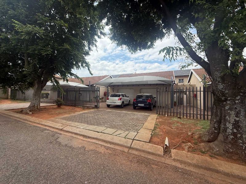 Front exterior view of a townhouse featuring a double garage, paved driveway, shadeport, and secure metal perimeter fencing.