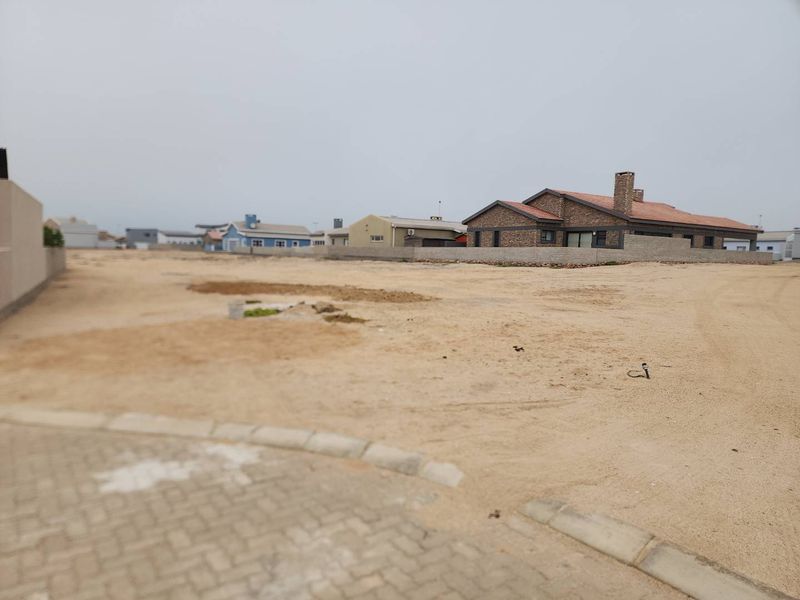 A vacant residential plot featuring sandy terrain, a paved curb, and surrounding boundary walls in a developed neighborhood.