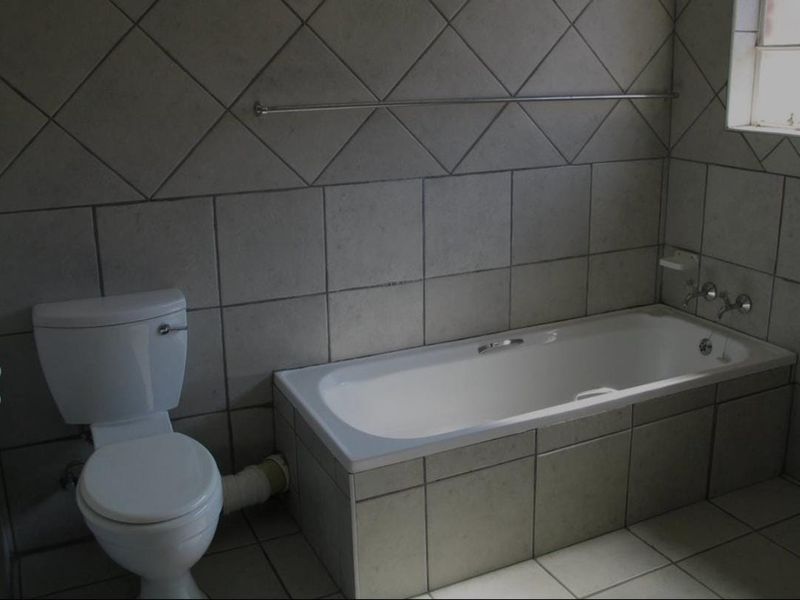 A dated bathroom featuring tiled walls and flooring, a built-in bathtub, and a toilet, requiring cosmetic updates and plumbing concealment.