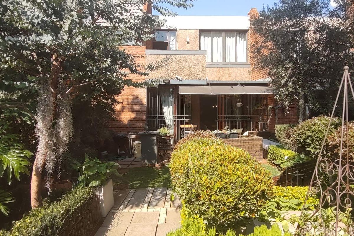 Rear view of a townhouse featuring a brick facade, a paved patio area with a retractable awning, and a well-established landscaped garden.