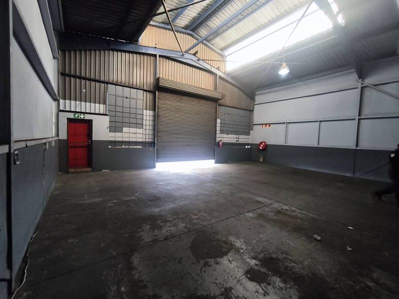 Spacious industrial warehouse unit featuring high ceilings, a large roller shutter door, concrete flooring, and a dedicated fire exit.