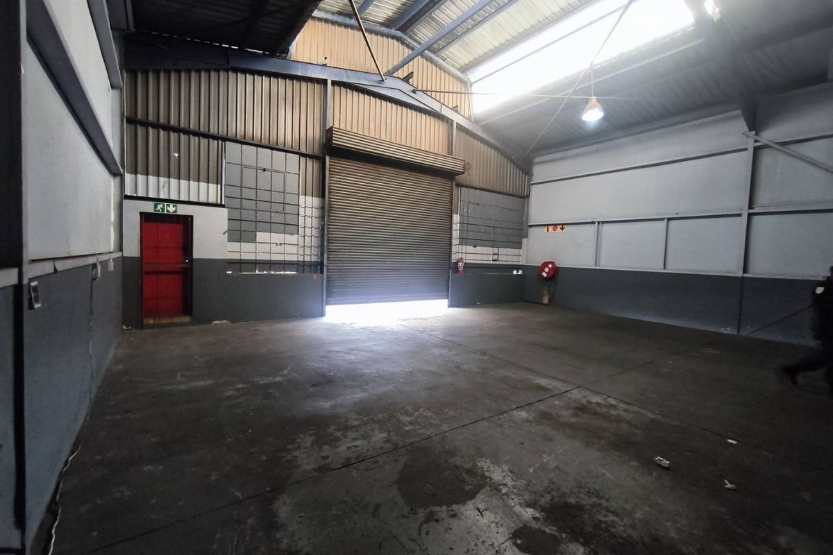 Spacious industrial warehouse unit featuring high ceilings, a large roller shutter door, concrete flooring, and a dedicated fire exit.