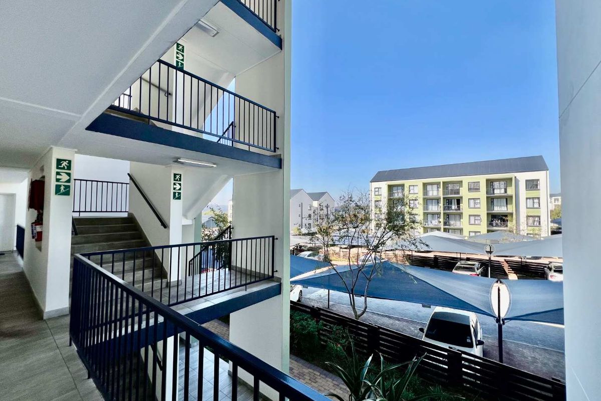 View of the apartment complex exterior, featuring open-air stairwells with metal railings and a view of the parking area with shade cloth structures.