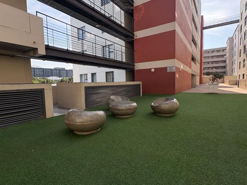 An open-air communal terrace area featuring artificial turf, metal safety railings, and an elevated walkway connecting the apartment building sections.