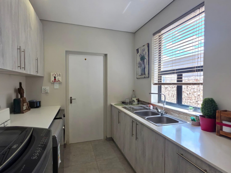 A functional scullery featuring modern built-in cabinetry, stone countertops, a double basin sink, and tiled flooring with natural light from a window.