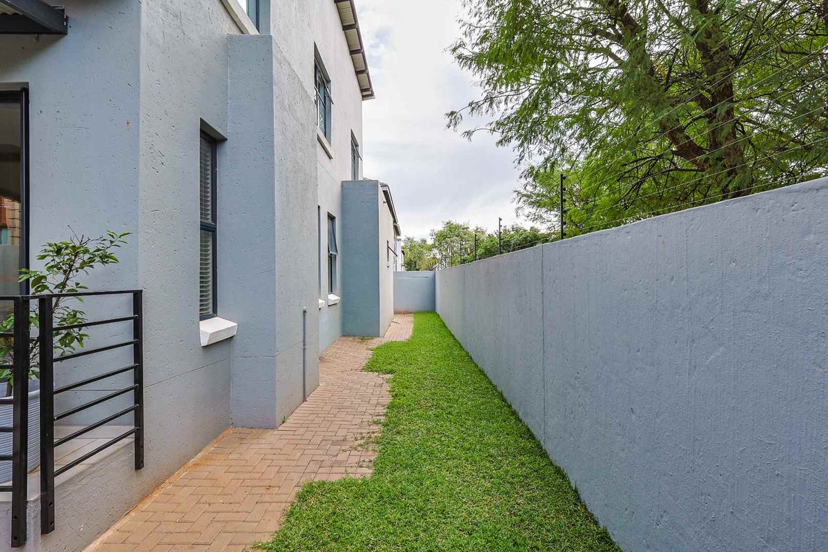 A side exterior view of the property featuring a paved walkway, a small grass strip, and a high boundary wall with electric fencing.