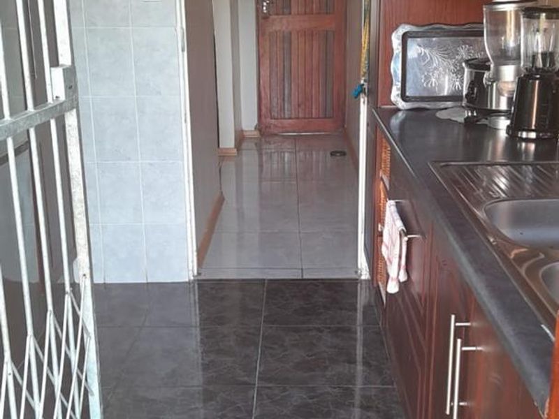 A functional scullery area featuring dark wood cabinetry, laminate countertops, a stainless steel sink, and tiled flooring, leading into the main hallway.