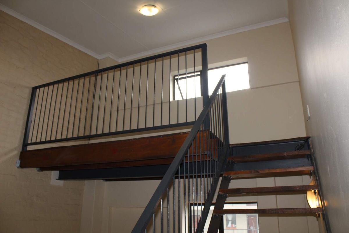 An open-plan living area featuring a mezzanine level accessed by a metal staircase with timber treads and exposed brick wall detailing.