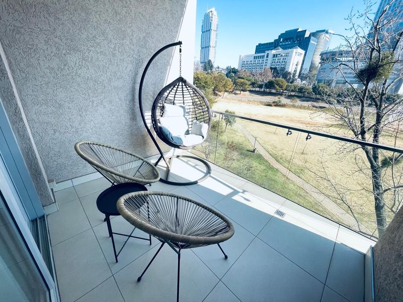A modern balcony featuring a glass balustrade and tiled flooring, offering expansive views of the surrounding urban landscape and parkland.