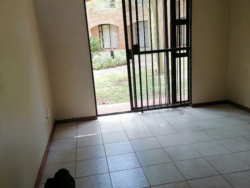 An empty living room featuring tiled flooring and a sliding glass door with a security gate, providing direct access to the outdoor garden area.